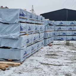 Stosy tarcicy zabezpieczone folią z logo ATA Timber Sweden, ułożone na zewnątrz w zimowej scenerii, z widocznymi elementami konstrukcyjnymi i liniami wysokiego napięcia w tle.