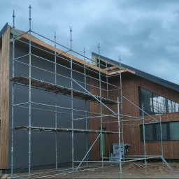 INDUSTRY DESIGN MACIEJ PUTYRA - Nowoczesny dom z drewnianą elewacją i szarymi panelami, w trakcie budowy. Widoczne rusztowania, okna o nietypowym kształcie i brama garażowa. Pochmurne niebo w tle.