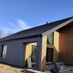 INDUSTRY DESIGN MACIEJ PUTYRA - Nowoczesny dom z elementami drewnianymi i szarą elewacją, widoczny z zewnątrz w słoneczny dzień. Uwagę zwraca pionowa boazeria i duże okna.
