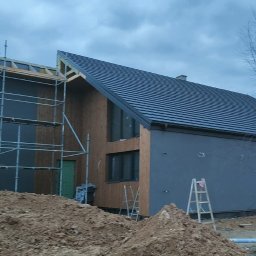 INDUSTRY DESIGN MACIEJ PUTYRA - Dom szkieletowy w trakcie budowy: widoczne rusztowania, fragmenty elewacji z drewna i szarego tynku, ciemna dachówka. Na pierwszym planie hałda ziemi.