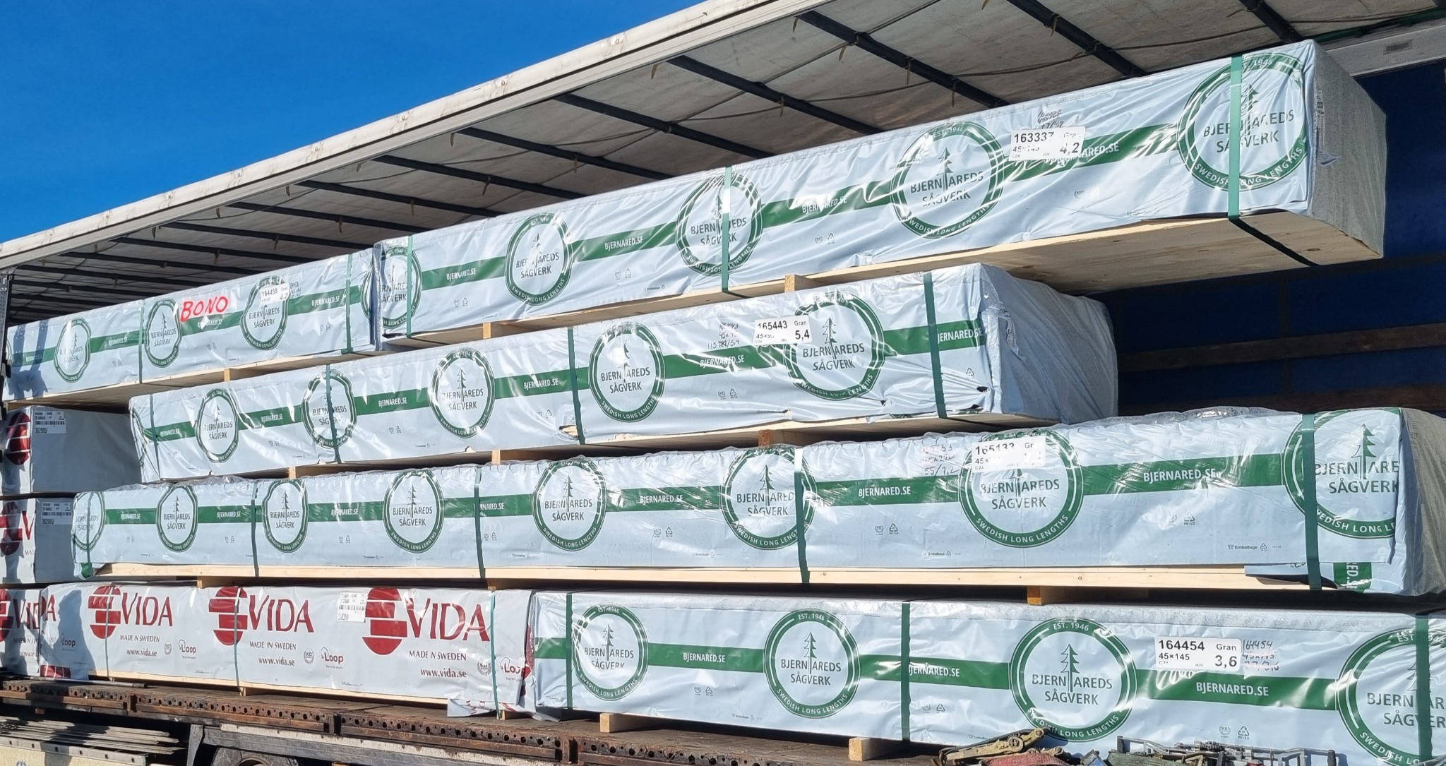 Załadowany na ciężarówkę pakiet tarcicy budowlanej w folii z logo producenta, widoczne oznaczenia specyfikacji i certyfikaty.