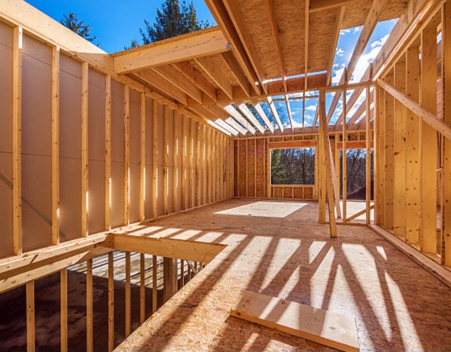 Konstrukcja szkieletowa domu w trakcie budowy, widoczne drewniane belki i płyty, niebo w tle przez otwory w dachu.