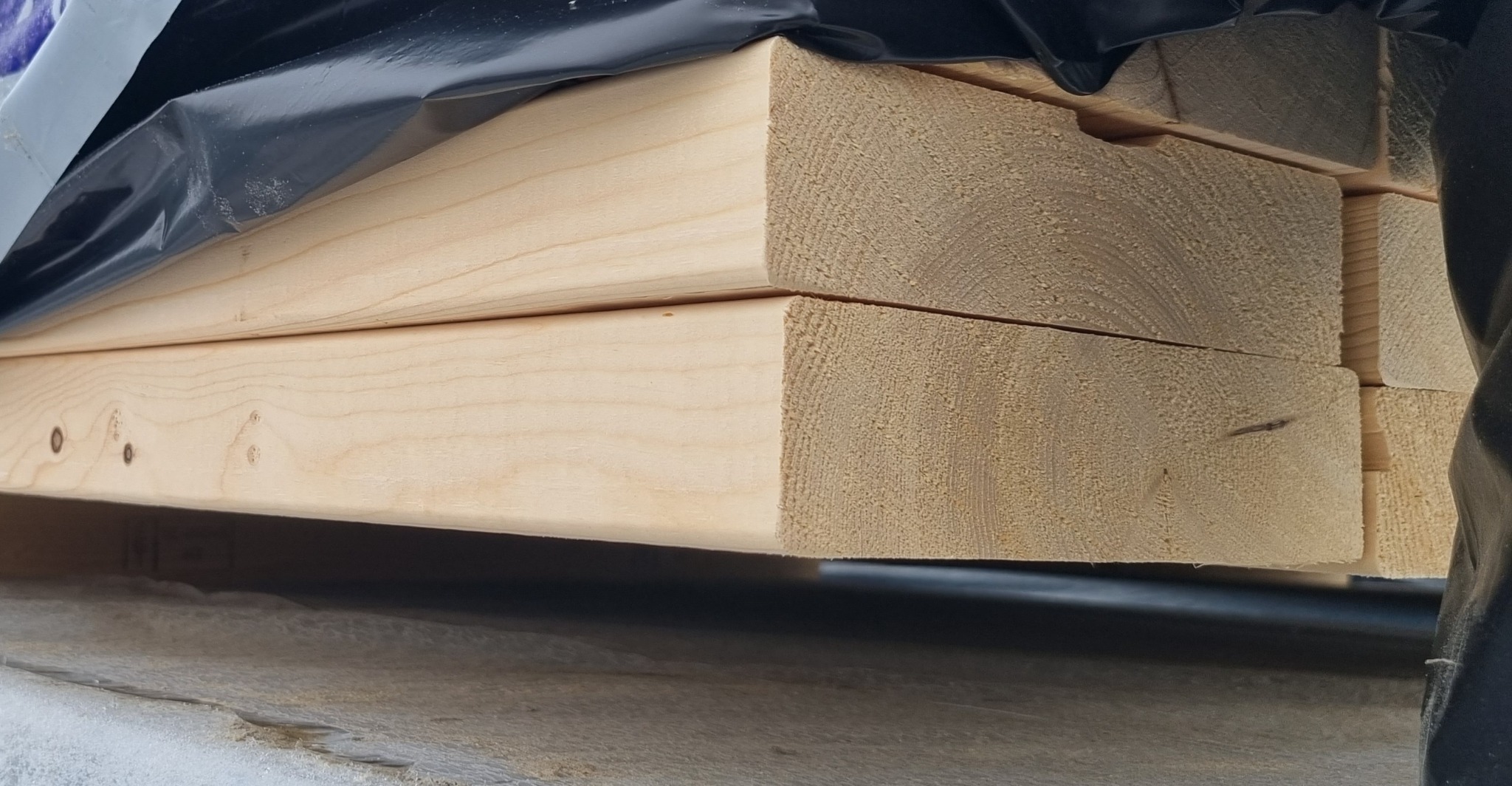 Stos jasnych, świeżo przetartych desek pod czarną folią ochronną, widoczne słoje drewna.