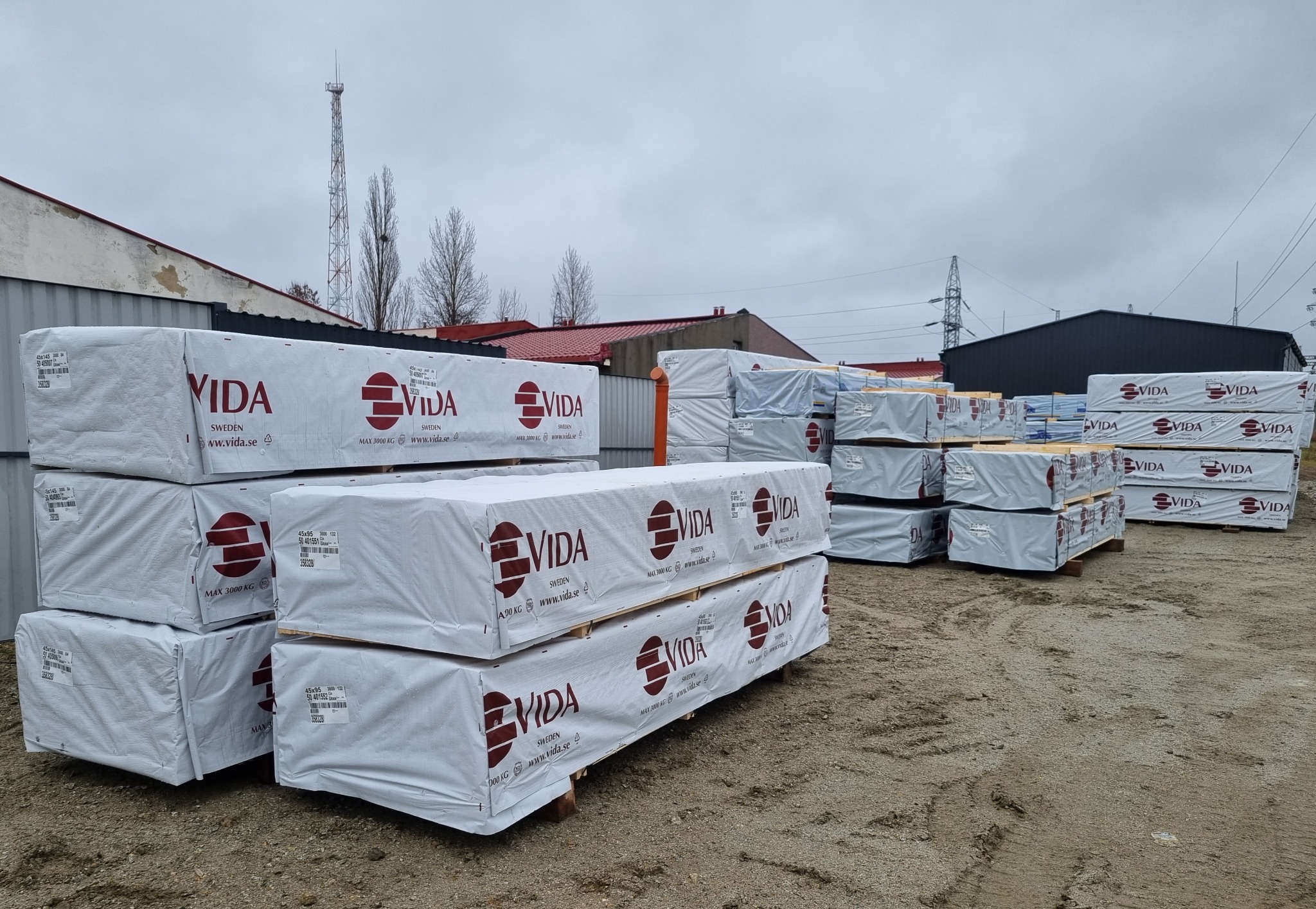 Sztaple zapakowanego drewna VIDA na placu składowym, widoczne budynki przemysłowe i maszt telekomunikacyjny na pochmurnym niebie.