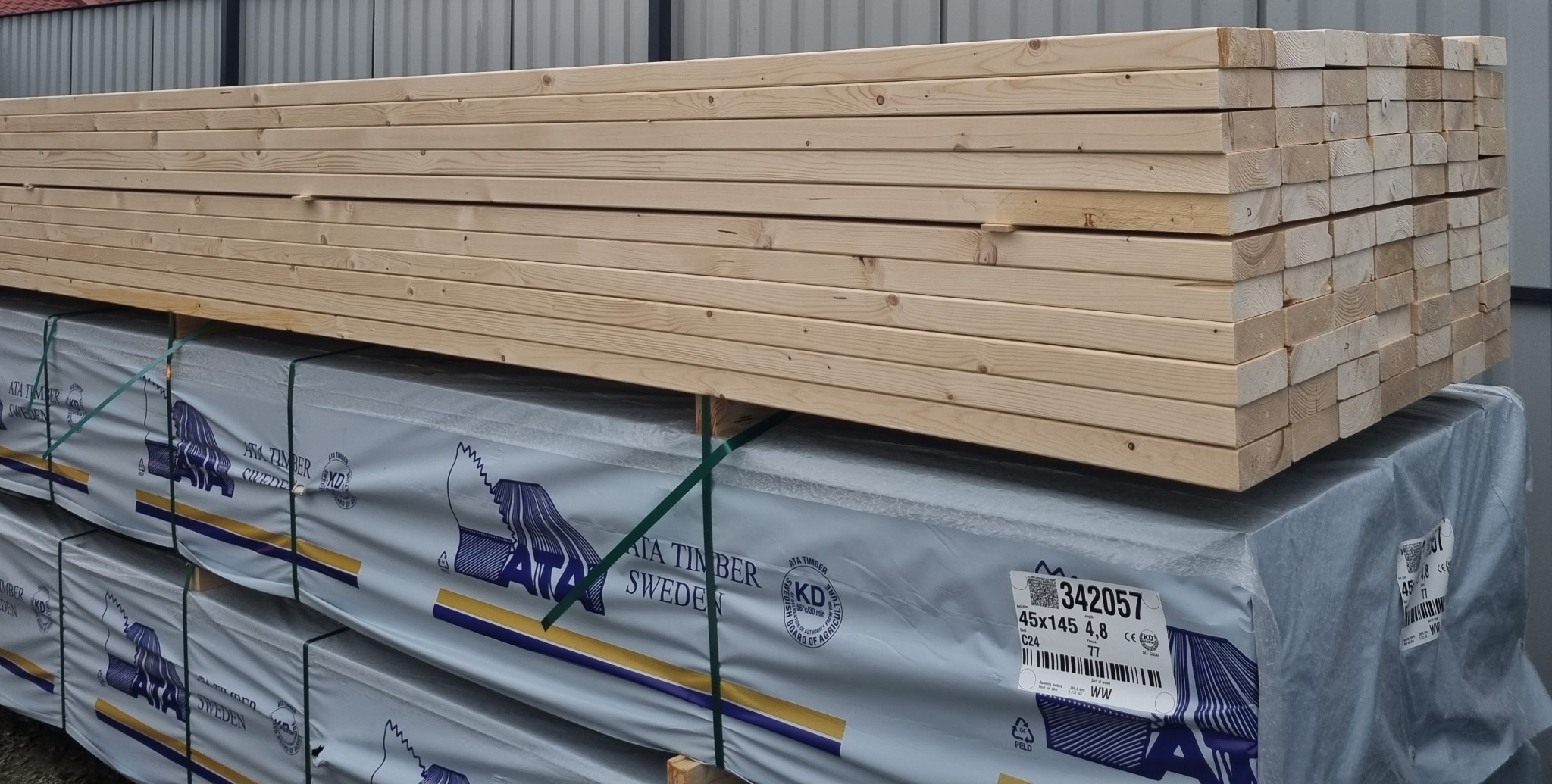 Stos świeżego drewna konstrukcyjnego na palecie, zabezpieczony folią z logo producenta, widoczne oznaczenia norm i certyfikatów.