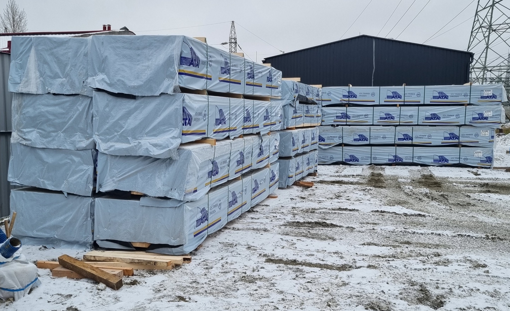 Stosy tarcicy zabezpieczone folią z logo ATA Timber Sweden, ułożone na zewnątrz w zimowej scenerii, z widocznymi elementami konstrukcyjnymi i liniami wysokiego napięcia w tle.