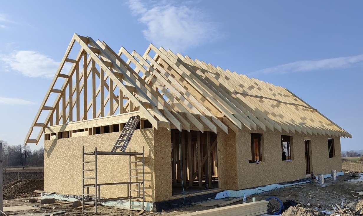 Wznoszenie domu szkieletowego: konstrukcja ścian z płyt OSB i drewniany szkielet dachu w słoneczny dzień. Widoczne rusztowania i drabina.
