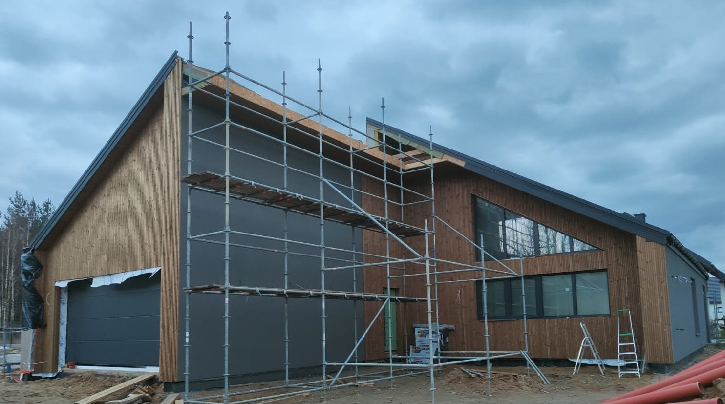 Nowoczesny dom z drewnianą elewacją i szarymi panelami, w trakcie budowy. Widoczne rusztowania, okna o nietypowym kształcie i brama garażowa. Pochmurne niebo w tle.
