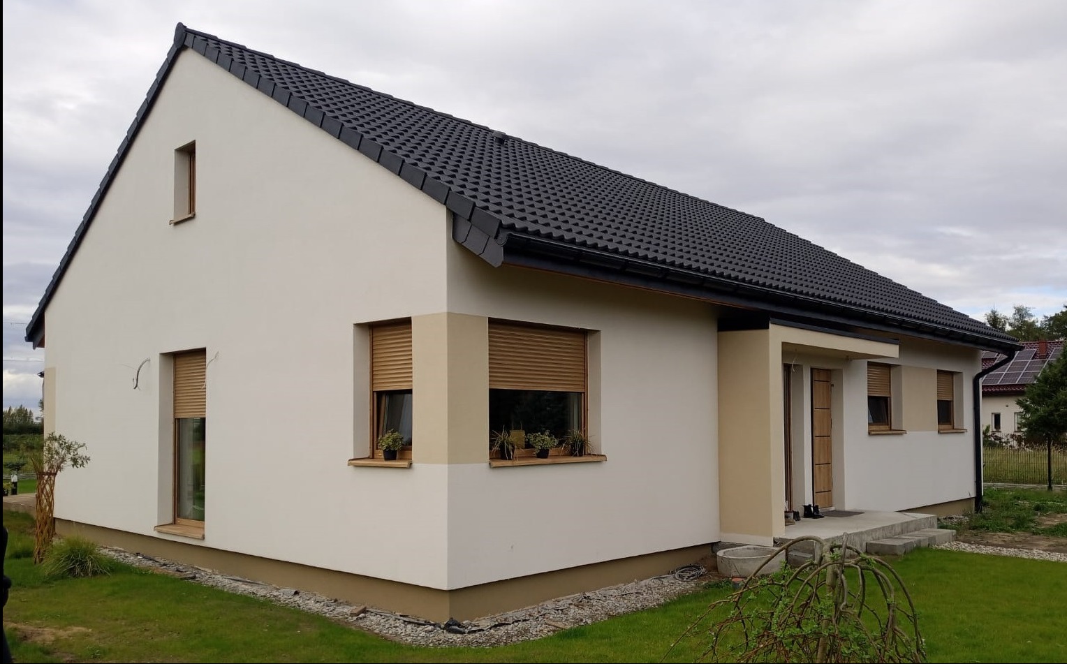 Nowoczesny, parterowy dom z ciemną dachówką i jasną elewacją. Okna z roletami zewnętrznymi i kwiatami na parapetach. Widok z boku na zadbany trawnik i fragment ogrodu.
