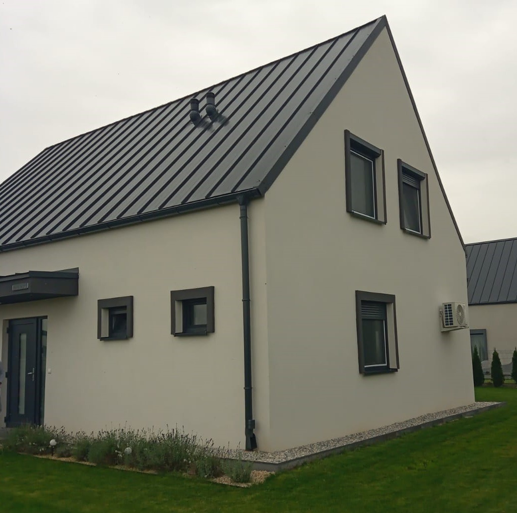 Nowoczesny dom z jasną elewacją i ciemnym dachem z blachy na tle pochmurnego nieba. Widoczne małe okna z ciemnymi ramami i klimatyzator na ścianie.
