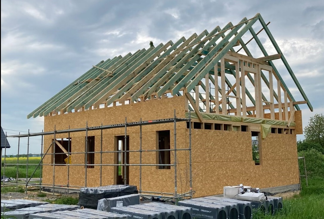 Budowa domu szkieletowego w trakcie: ściany z płyt OSB, drewniana konstrukcja dachu z zielonymi krokwiami, rusztowania. Na pierwszym planie materiały budowlane.