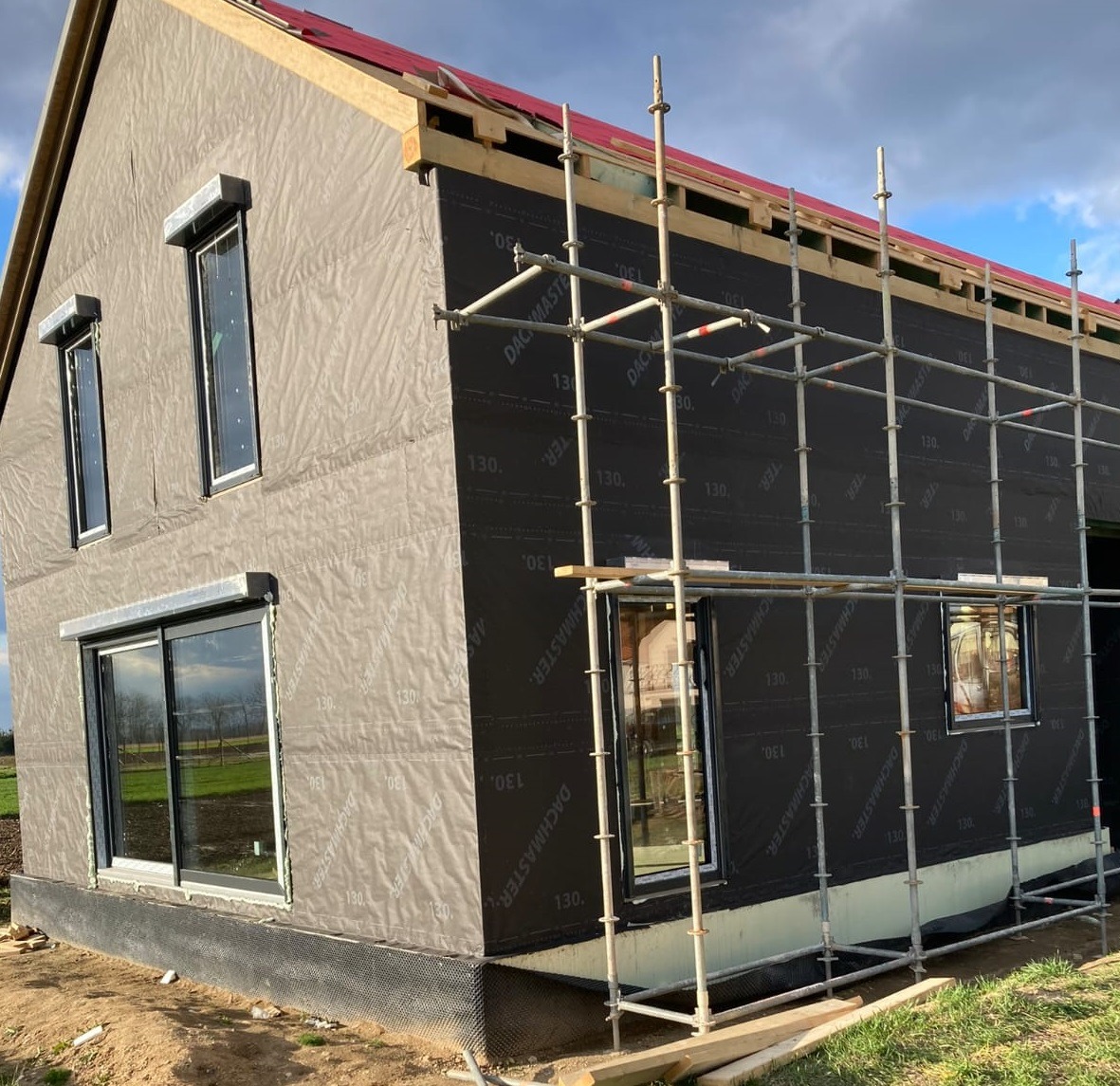 Budowa domu w trakcie: widoczne okna, rusztowania i czarna membrana dachowa. Częściowo wykończona elewacja. Ujęcie z zewnątrz, w słoneczny dzień.