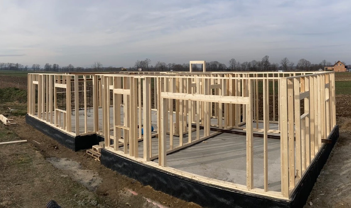 Szkielet domu w trakcie budowy, widoczne drewniane ramy ścian na fundamencie z izolacją, krajobraz polny w tle, pochmurne niebo.