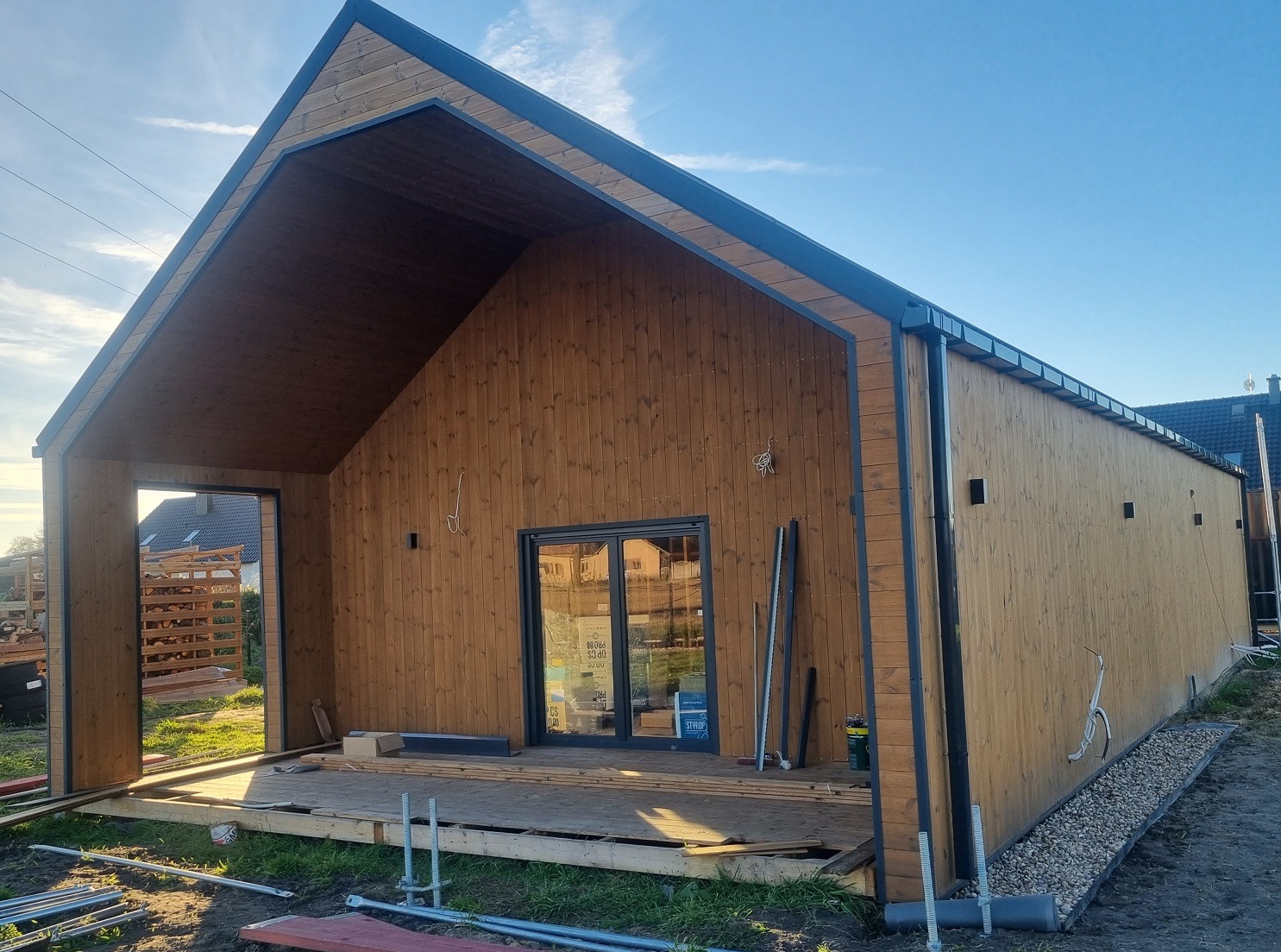 Surowy, brązowy dom z drewna w trakcie budowy, z widoczną konstrukcją dachu i elementami wykończeniowymi. Widoczne narzędzia i materiały budowlane.