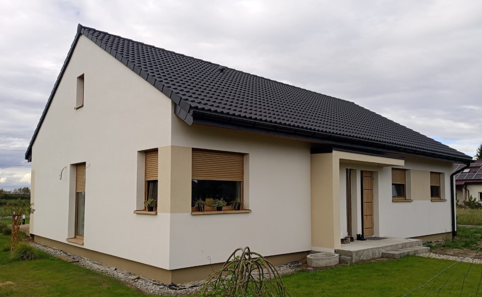 Parterowy dom z ciemną dachówką, jasną elewacją i roletami zewnętrznymi. Widok z boku na wejście i okna z roślinami. Ujęcie w pochmurny dzień.