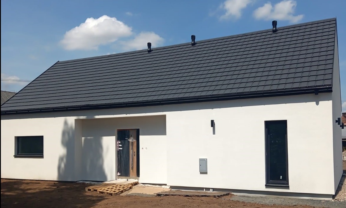 Nowoczesny, parterowy dom z ciemną dachówką i białymi ścianami. Widoczne okna, drzwi wejściowe zabezpieczone folią, palety przed wejściem. Błękitne niebo z chmurami w tle.