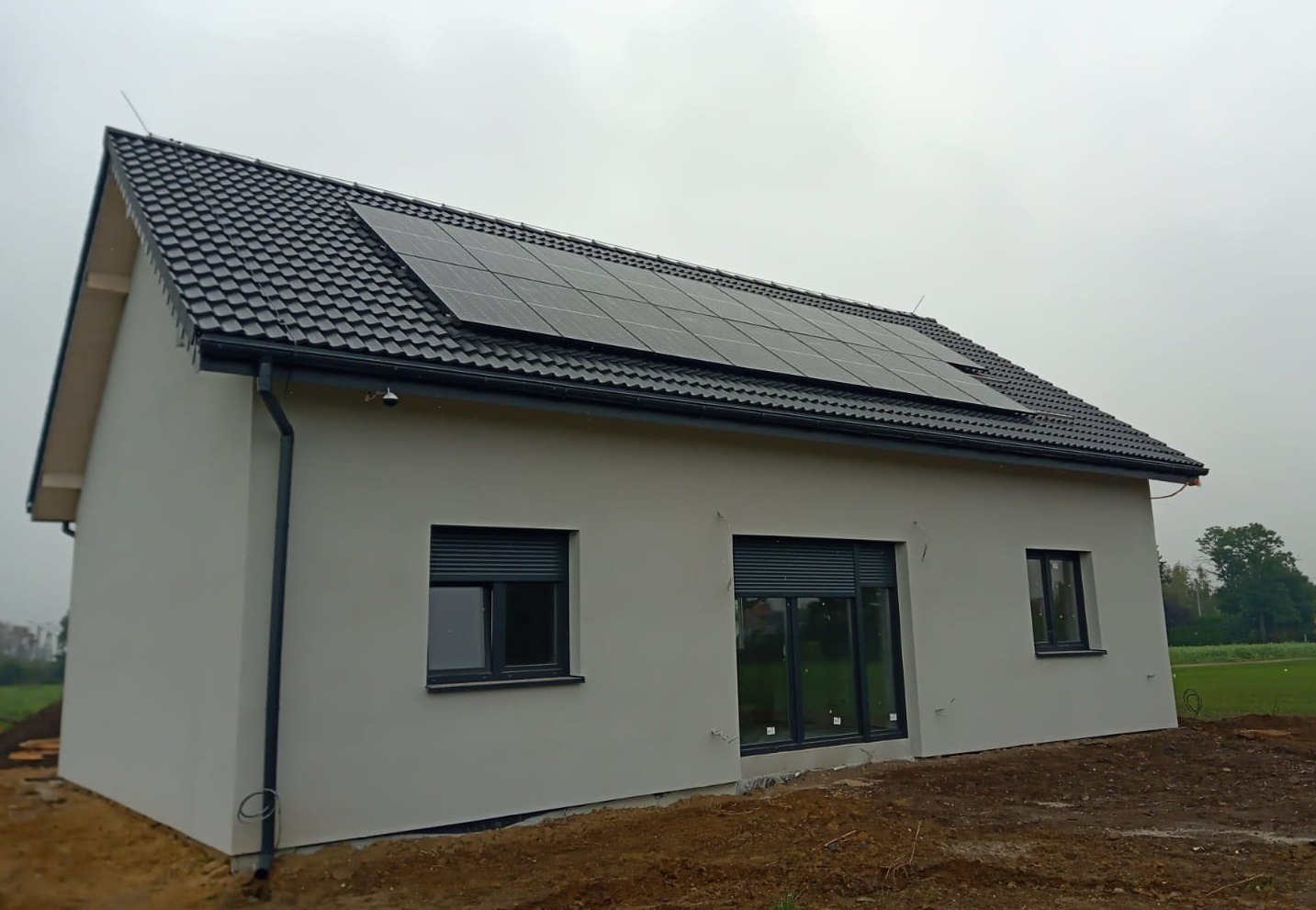 Gotowy, parterowy dom z panelami słonecznymi na dachu, szare okna z roletami zewnętrznymi, elewacja w jasnym odcieniu szarości, widok z boku na tle pochmurnego nieba.