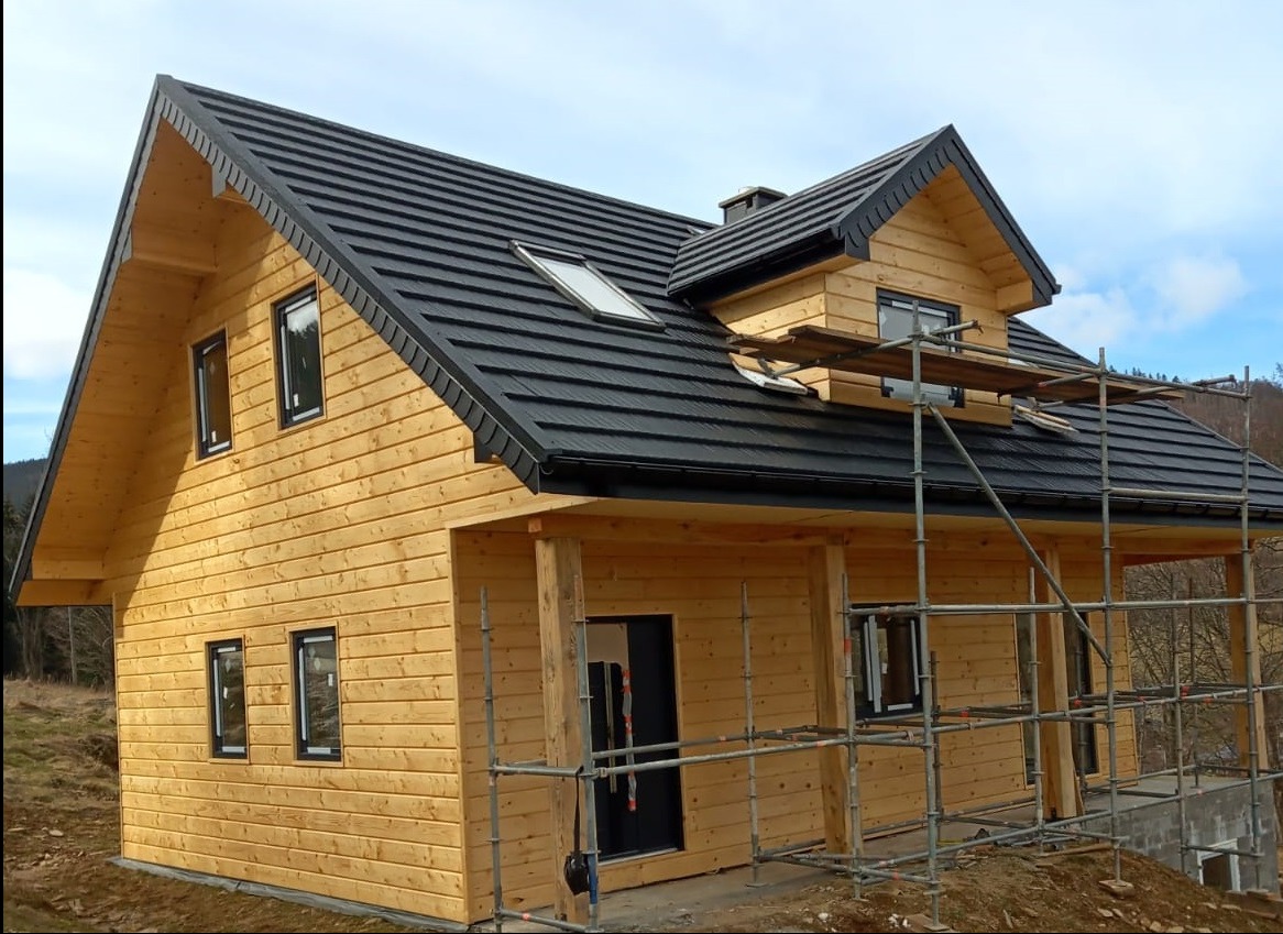 Surowy, dwukondygnacyjny dom z jasnego drewna z czarnym dachem, w trakcie budowy, otoczony rusztowaniami. Widoczne okna i drzwi. W tle zalesione wzgórza.
