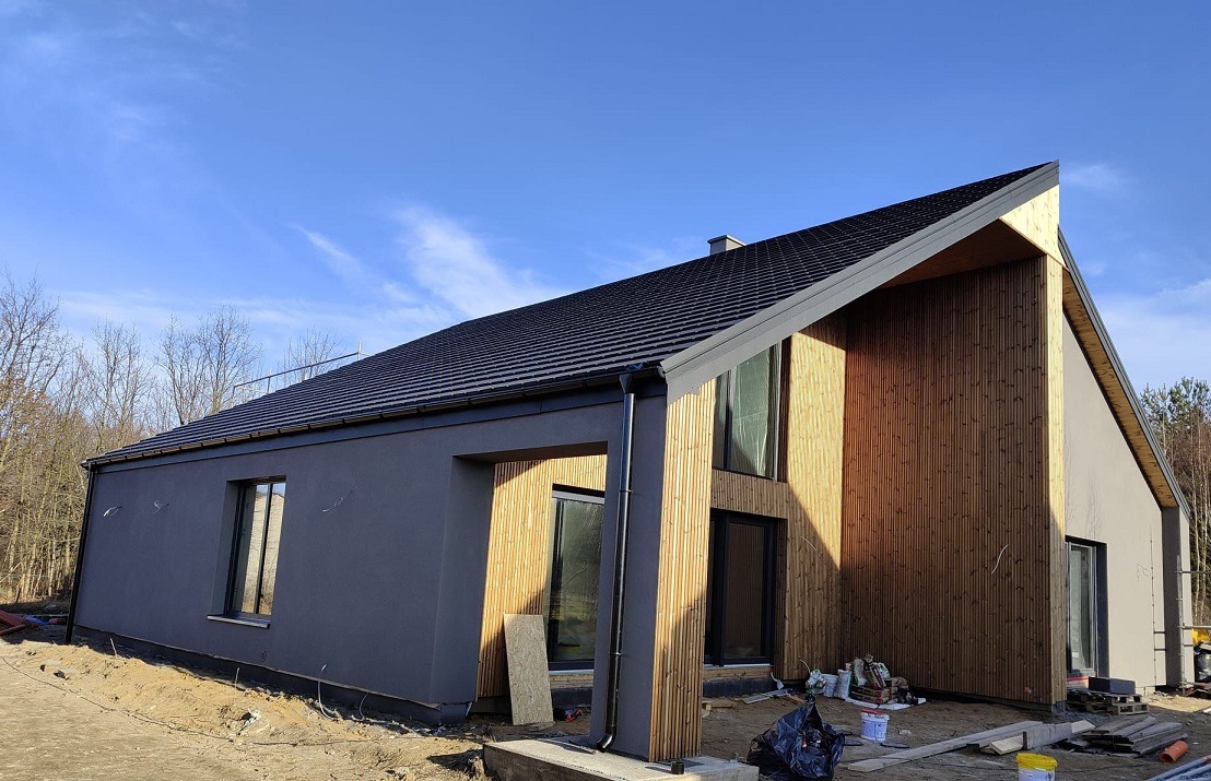 Nowoczesny dom z elementami drewnianymi i szarą elewacją, widoczny z zewnątrz w słoneczny dzień. Uwagę zwraca pionowa boazeria i duże okna.