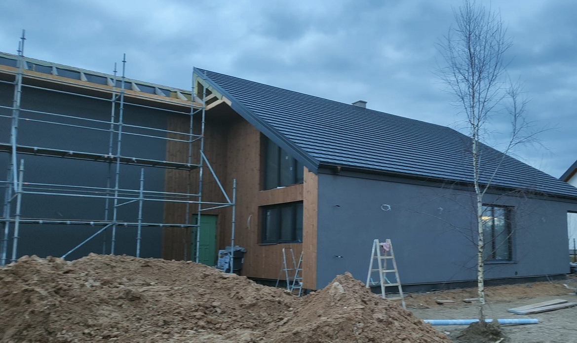 Dom szkieletowy w trakcie budowy: widoczne rusztowania, fragmenty elewacji z drewna i szarego tynku, ciemna dachówka. Na pierwszym planie hałda ziemi.
