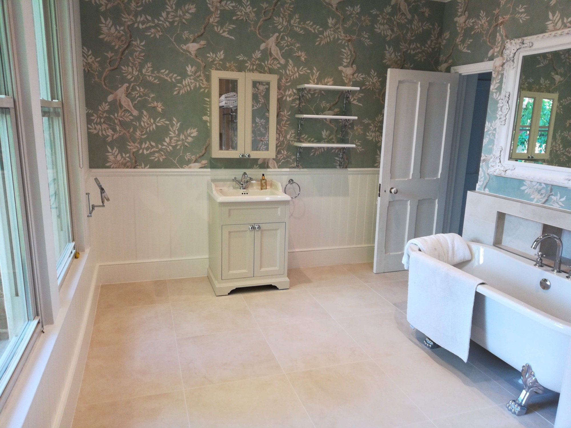Jasna łazienka z klasyczną wanną na lwich nóżkach, umywalką z szafką i lustrem, zieloną tapetą w ptaki i białymi panelami ściennymi.