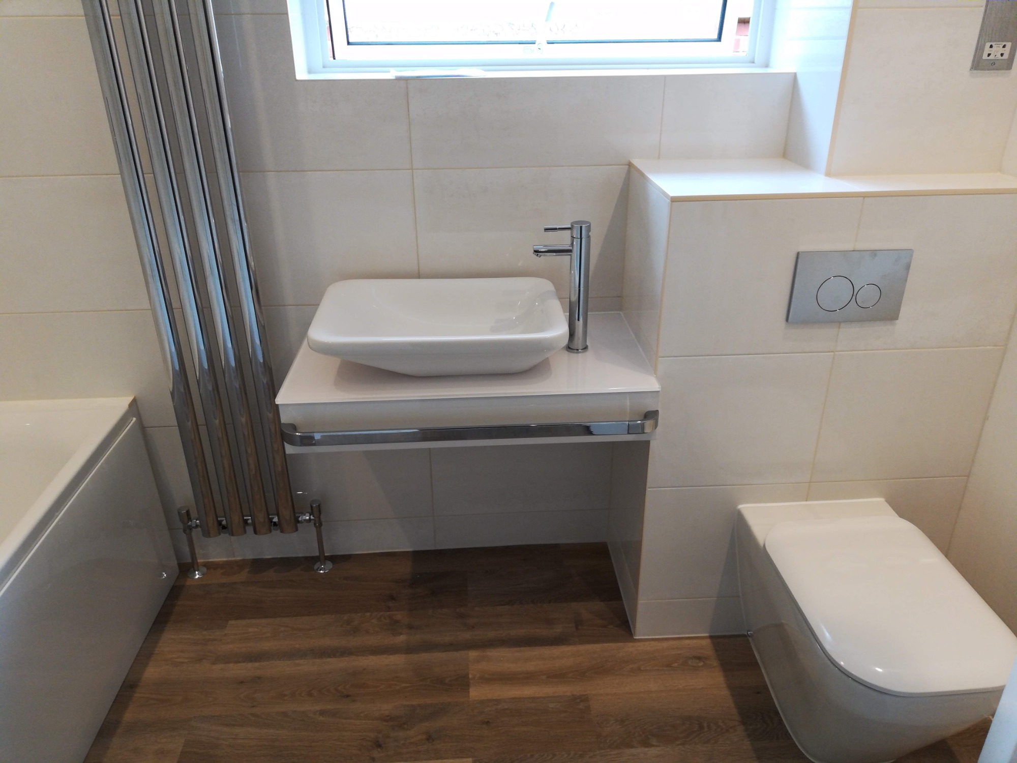 Nowoczesna łazienka z beżowymi płytkami, kwadratową umywalką nablatową, chromowaną armaturą i grzejnikiem drabinkowym, drewnopodobna podłoga, wisząca toaleta z przyciskiem spłukującym w metalowej...