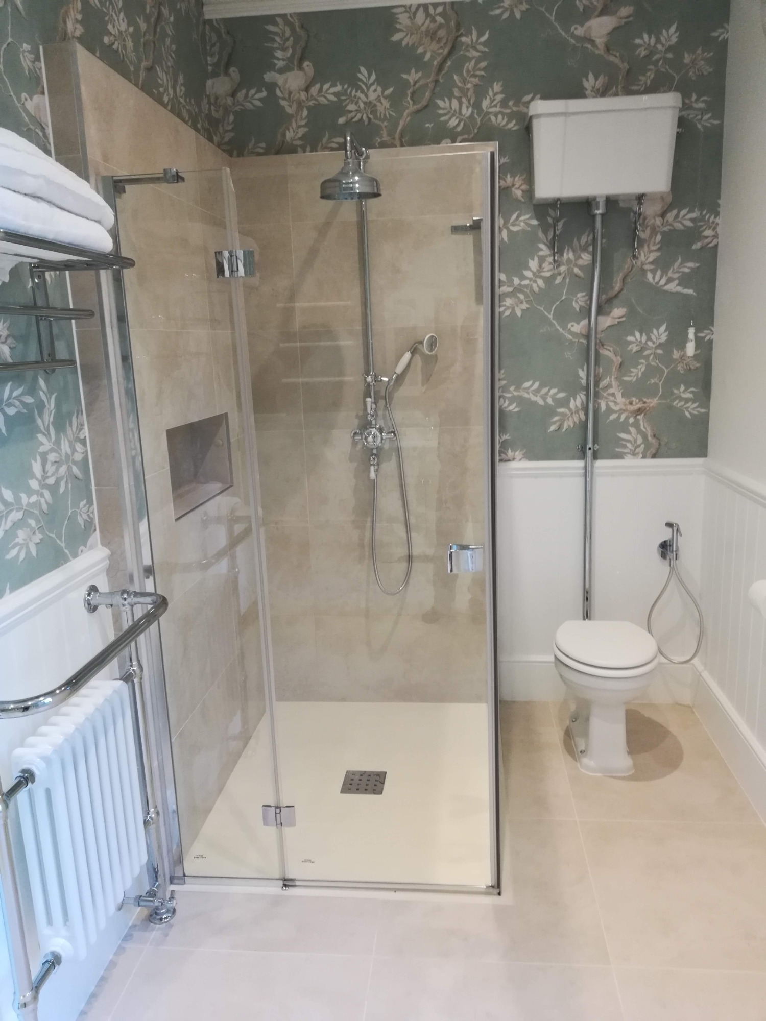 Nowoczesna łazienka z kabiną prysznicową typu walk-in, beżowymi płytkami, toaletą w stylu retro i grzejnikiem drabinkowym, na ścianie tapeta w ptaki i gałęzie.