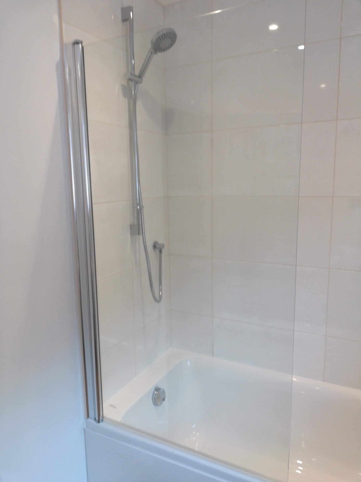 Biała wanna z zamontowanym szklanym parawanem i chromowaną baterią prysznicową na tle jasnych, kwadratowych płytek.