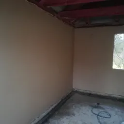 Świeżo otynkowane ściany w surowym wnętrzu z widokiem na zewnątrz przez okno, widoczne czerwone elementy konstrukcji dachu.