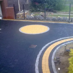 Ułożona nawierzchnia z ciemnej kostki brukowej z dekoracyjnym okręgiem z żółtej kostki, obrzeżem z szarej kostki granitowej i paletą kostki w tle.