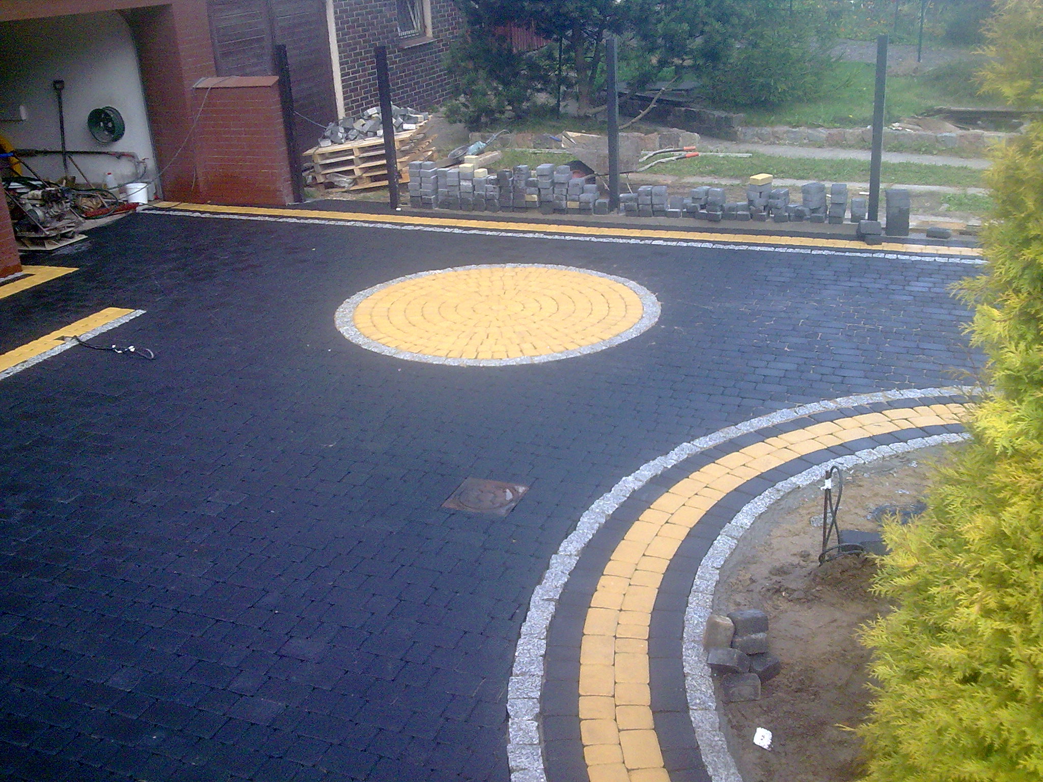 Ułożona nawierzchnia z ciemnej kostki brukowej z dekoracyjnym okręgiem z żółtej kostki, obrzeżem z szarej kostki granitowej i paletą kostki w tle.