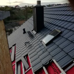 Częściowo pokryty grafitowy dach z kominem, oknem dachowym i elementami montażowymi, widoczna czerwona membrana dachowa i drewniana konstrukcja więźby.