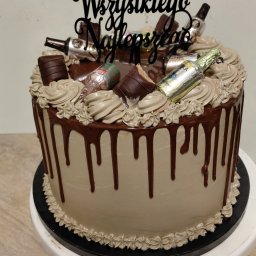 Przetwórnia Smaku Arleta Olbrych - Tort urodzinowy z czekoladowymi dekoracjami, mini buteleczkami i napisem 'Wszystkiego Najlepszego' na białym stojaku. Kremowy tort z polewą.