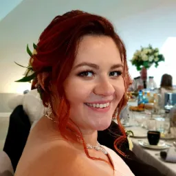 Uśmiechnięta, ruda kobieta z kwiatami we włosach na tle stołu weselnego z gośćmi w tle. Widoczne napoje i jedzenie.