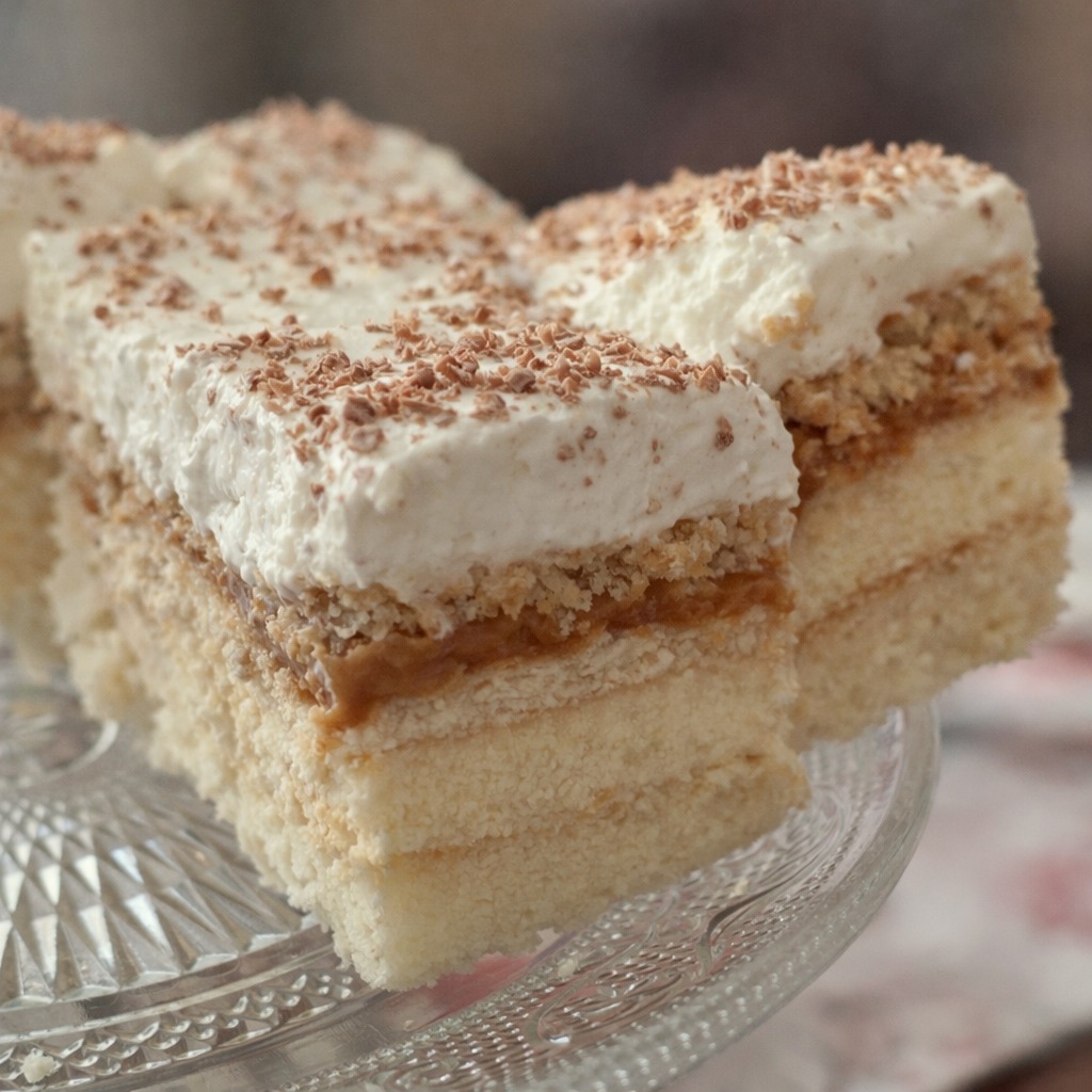 Kawałek kremowego ciasta z posypką czekoladową na szklanym talerzu. Widoczne warstwy biszkoptu, kremu i karmelu. Delikatne oświetlenie podkreśla teksturę.