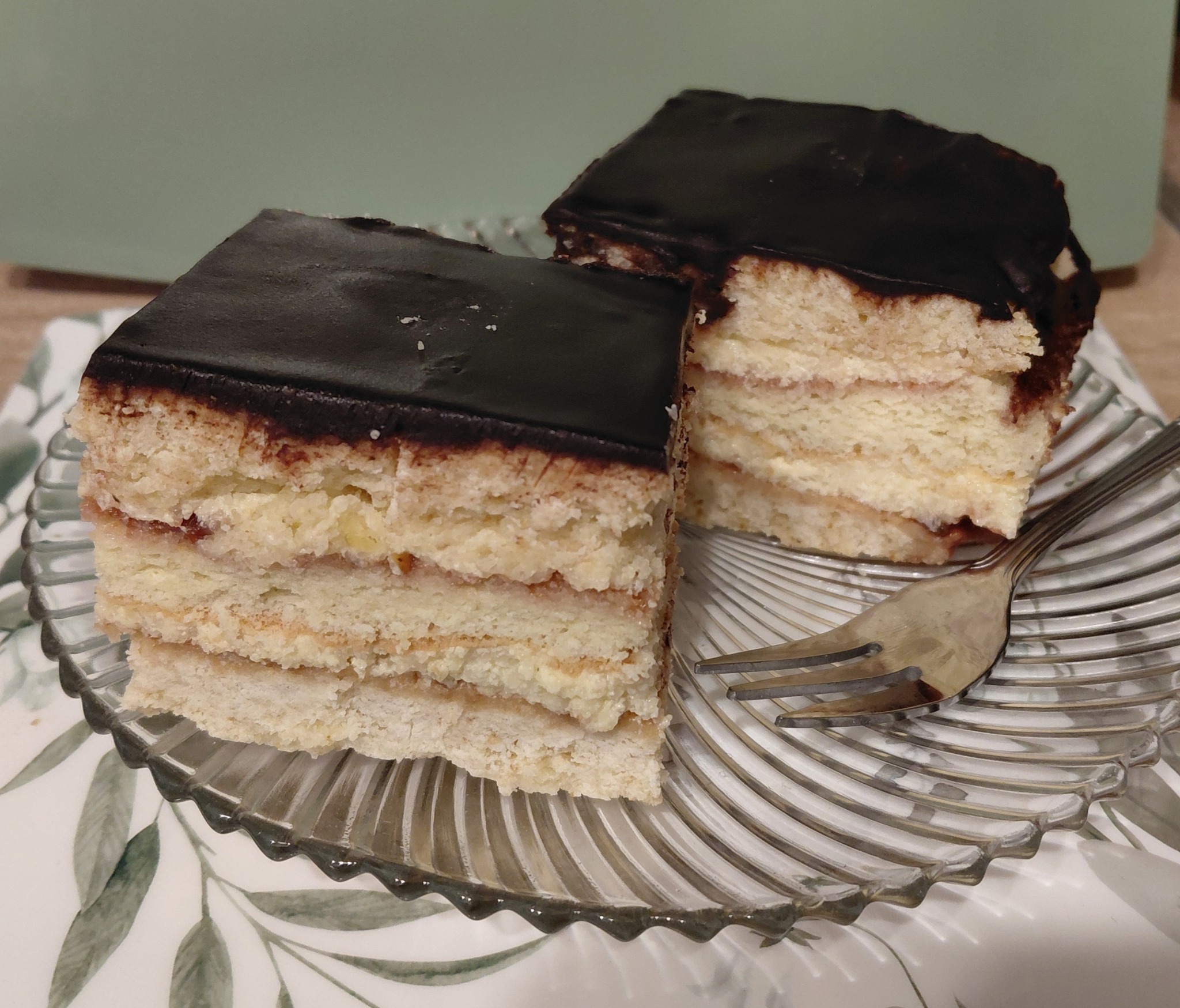 Dwa kawałki tortu z polewą czekoladową na szklanym talerzu z widelcem. Tło w odcieniach zieleni i beżu. Widoczne warstwy ciasta i kremu.