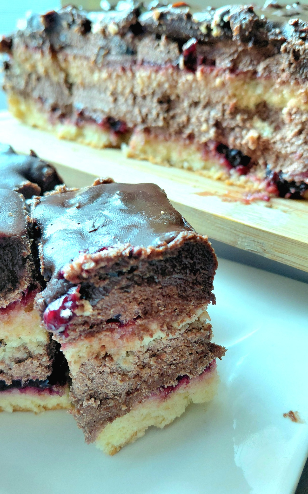 Kawałek tortu z wiśniami i czekoladową polewą na białym talerzu, z widocznym przekrojem warstw ciasta i kremu. W tle reszta tortu na desce.