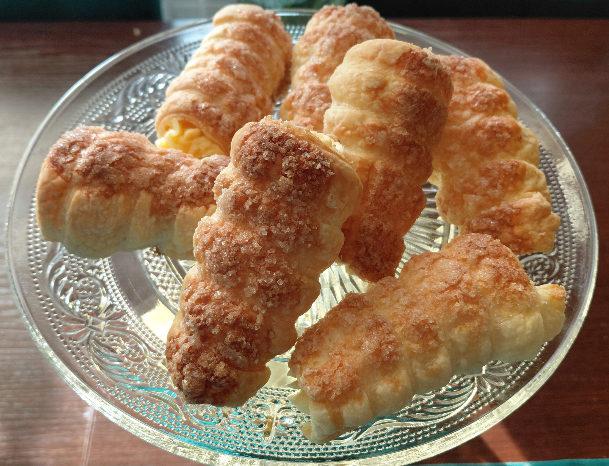 Kremówki w kształcie rożków, posypane cukrem, ułożone na szklanym, zdobionym talerzu. Widoczne kremowe nadzienie. Słodki deser.