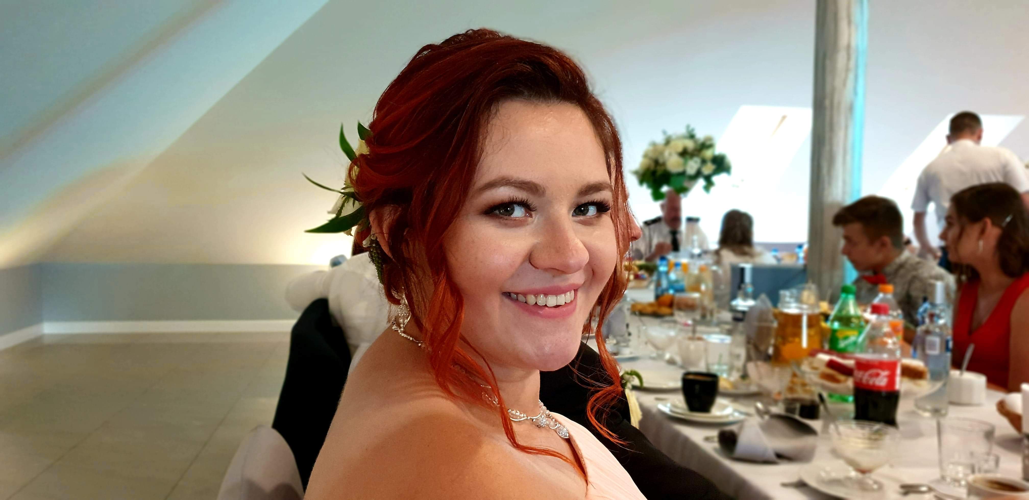 Uśmiechnięta, ruda kobieta z kwiatami we włosach na tle stołu weselnego z gośćmi w tle. Widoczne napoje i jedzenie.