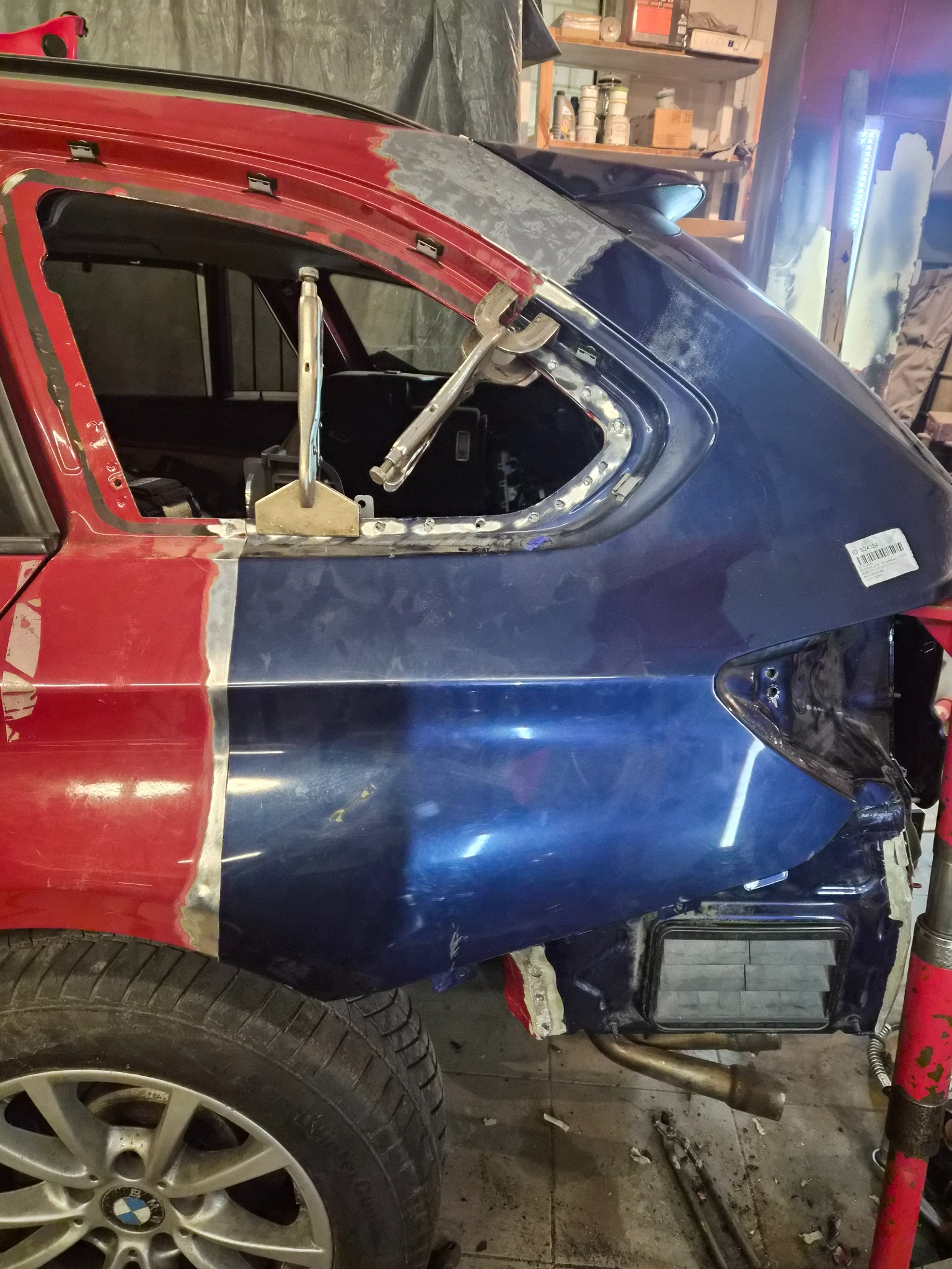 Częściowo naprawiony, czerwono-niebieski samochód BMW w warsztacie. Widoczne narzędzia i spawane elementy karoserii. Prace blacharsko-lakiernicze w toku.
