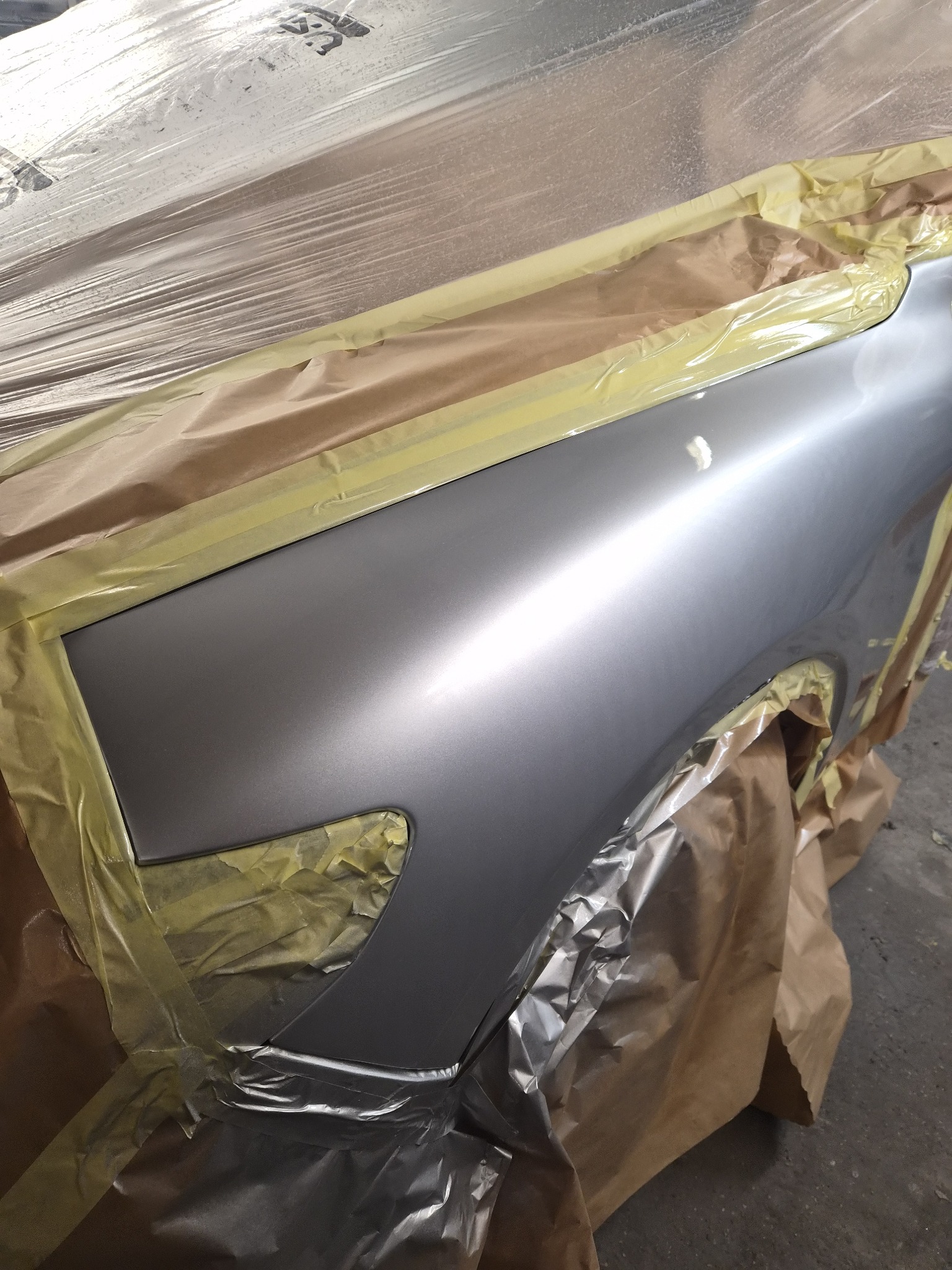 Szary błyszczący błotnik samochodu, zabezpieczony taśmą i folią malarską, przygotowany do lakierowania. Widoczne detale zabezpieczeń i świeżo położona warstwa lakieru.