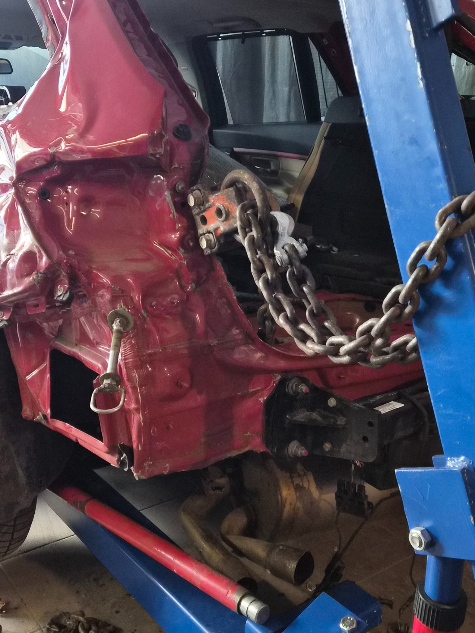 Uszkodzony, czerwony samochód na podnośniku warsztatowym, zabezpieczony łańcuchami. Widoczne zniekształcenia karoserii i elementy podwozia. Detal naprawy blacharskiej.