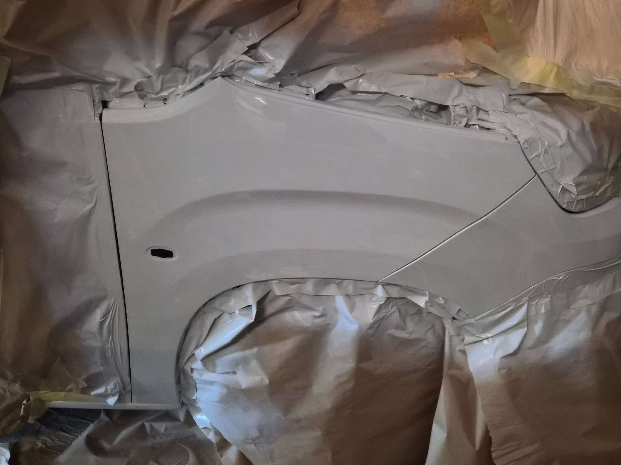 Biały błotnik samochodu po lakierowaniu, zabezpieczony folią malarską. Widoczny fragment karoserii w trakcie prac lakierniczych.