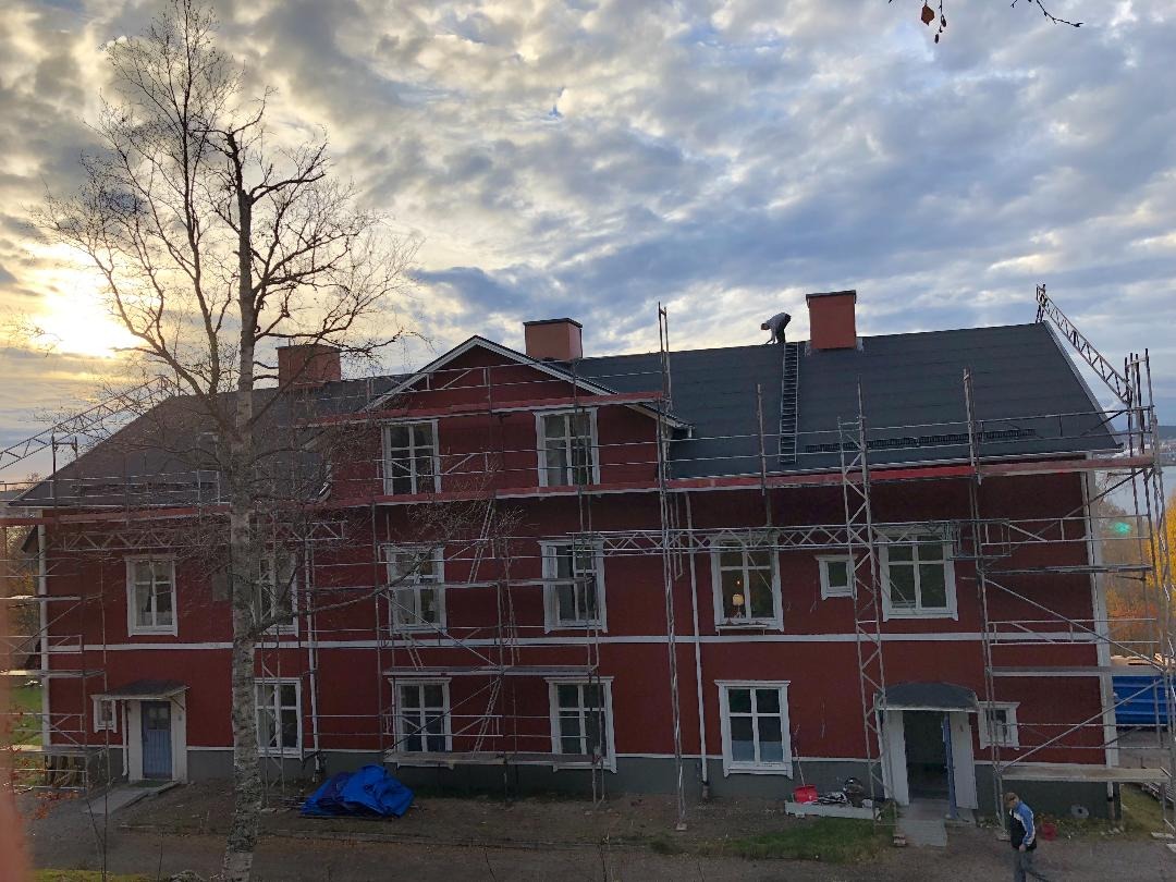 Czerwony, dwupiętrowy budynek mieszkalny w trakcie prac elewacyjnych i dekarskich; widoczne rusztowania, częściowo nowy dach i pracownik na dachu, błękitny brezent na ziemi.