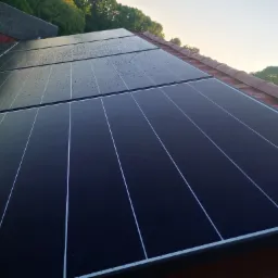 Panel słoneczny z kroplami deszczu na czerwonym dachu, widok z góry na tle drzew i budynku.