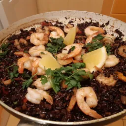 Paella z czarnym ryżem, krewetkami, cytryną i pietruszką, serwowana w dużej patelni z widocznymi przypaleniami na dnie.