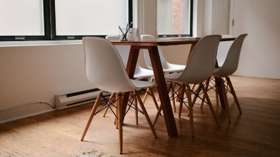 Jasne wnętrze z drewnianą podłogą, stół z ciemnego drewna i cztery białe krzesła z drewnianymi nogami, okno z białymi roletami w tle.