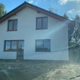 Nowo otynkowany dom jednorodzinny z drewnianą stolarką okienną, widok zza szyby samochodu, teren wokół domu niezagospodarowany.