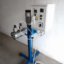 Stacjonarne urządzenie z metalowym cylindrem, silnikiem elektrycznym i panelem sterowania, osadzone na niebieskiej ramie z kółkami, widoczne kable i wtyczka.