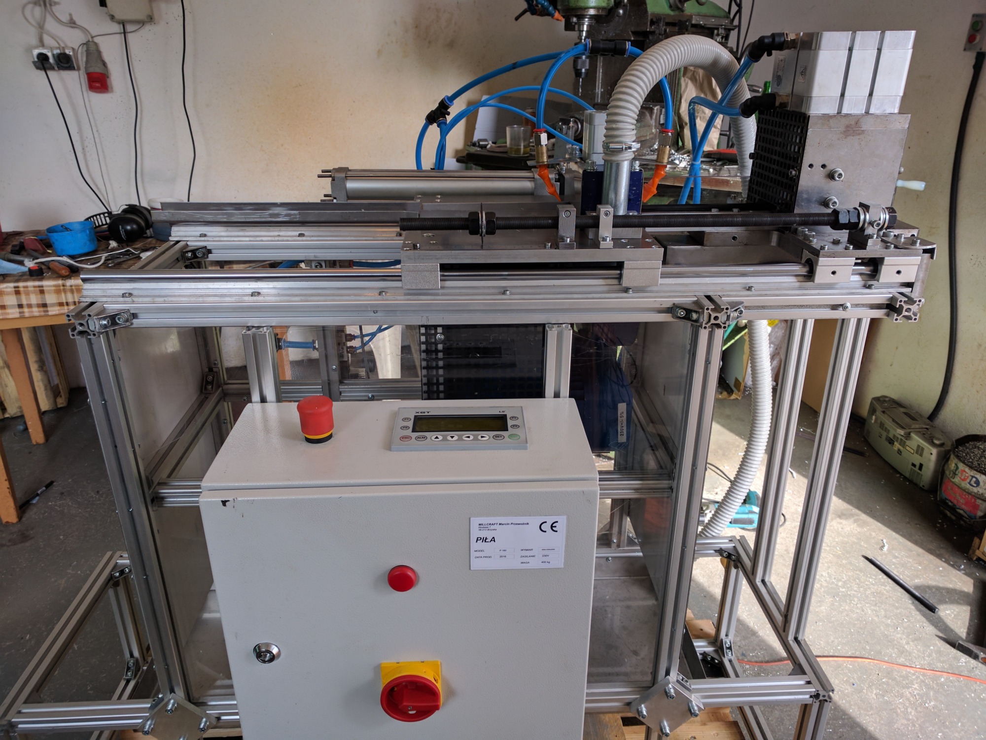 Widok z przodu na maszynę z aluminiową ramą, widoczne elementy sterujące, przewody pneumatyczne i fragment frezarki u góry.